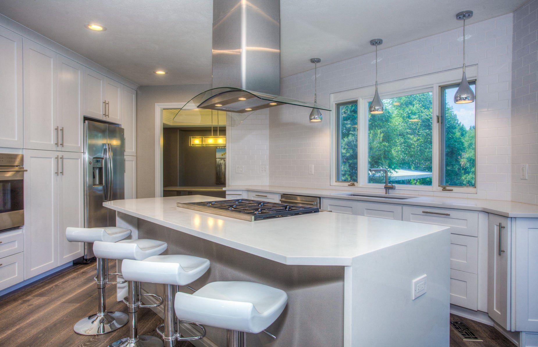 White kitchen with a gas cooktop in the island, a stainless side-by-side refrigerator with a water and ice dispenser, a wall oven at the far left and trees outside the windows